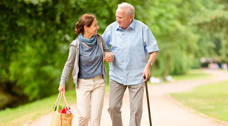 Eine Frau läuft mit einem älteren Herren durch einen Park und trägt seine Einkaufstasche