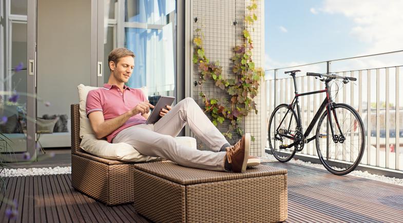 Ein Mann sitzt an einem sonnigen Tag auf dem Balkon und liest auf einem Tablet.