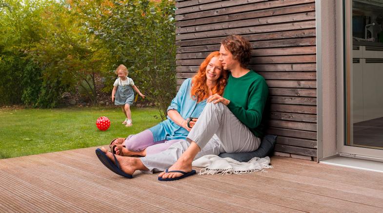Mann und Frau sitzen auf der Terrasse während die Tochter im Garten spielt.