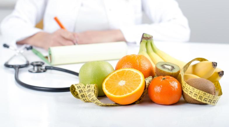 Arzt schreibt etwas, im Vorderund ist Obst und ein Maßband zu sehen